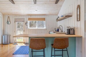 Une cuisine avec un plafond en bois, une fenêtre avec des stores, un évier, une machine à café et deux tabourets de bar.