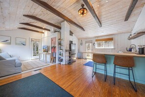 Een ruime woonkamer met een houten plafond, een bank, een salontafel, en een keuken met een bar en krukken.