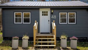 A small, dark gray building with a white door, numbered 48, and a wooden staircase leading to the entrance. - Romantic Forest Cabin - private, near everything (Bar Harbor)
