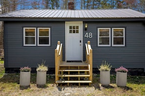 Un petit bâtiment de couleur gris foncé, avec une porte blanche, portant le numéro 48, et un escalier en bois menant à l’entrée.