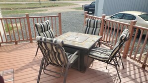 Une terrasse en bois avec une table et des chaises, une rambarde en métal, et une voiture garée à l’arrière-plan.