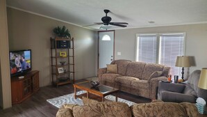 Un salon comprenant un canapé, une table basse, un téléviseur posé sur un meuble en bois, un ventilateur de plafond et une fenêtre avec des stores.