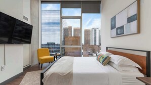 Un dormitorio moderno con una cama grande, una silla amarilla, un televisor y vistas a la ciudad a través de amplias ventanas.