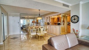 A spacious living area with a brown leather sofa, a dining table with chairs, a kitchen with wooden cabinets, and a wall-mounted clock. - Gorgeous 2/2 condo with fantastic views 1402 (Clearwater)