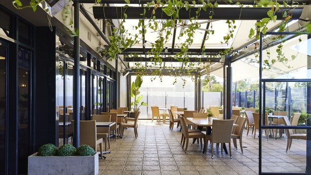 An outdoor café with tables and chairs, a pergola with hanging plants, and a view of the outdoors. - Blue Gums Hotel (Fairview Park)