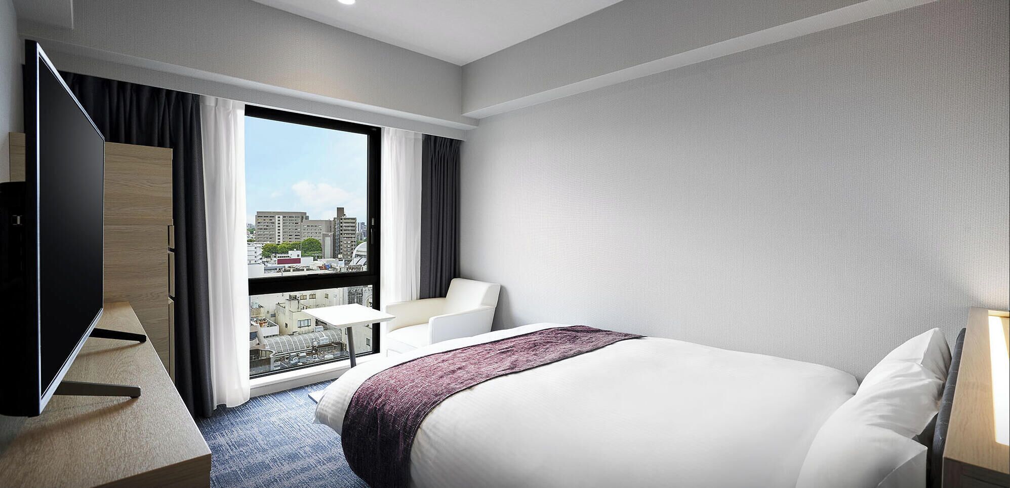A hotel room with a bed, a TV, a desk, a chair, and a window with a city view.