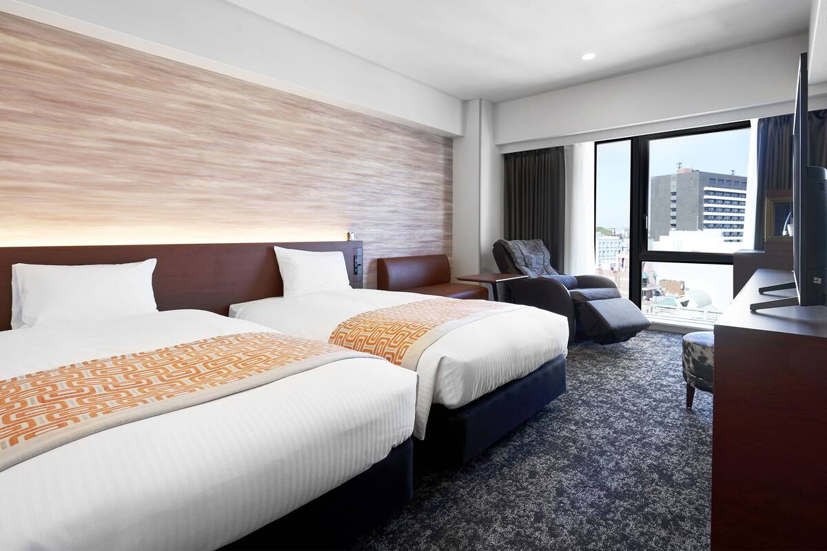 Habitación de hotel con dos camas, un escritorio, una silla y un ventanal con vista a la ciudad.