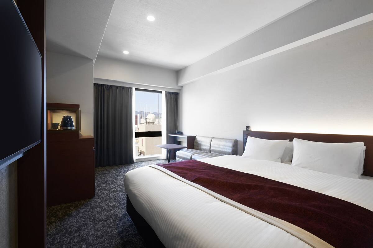 Habitación de hotel con una cama grande, un escritorio, una silla y vistas a la ciudad por la ventana.