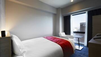 Quarto de hotel com cama, escrivaninha, cadeira e vista para a cidade pela janela.