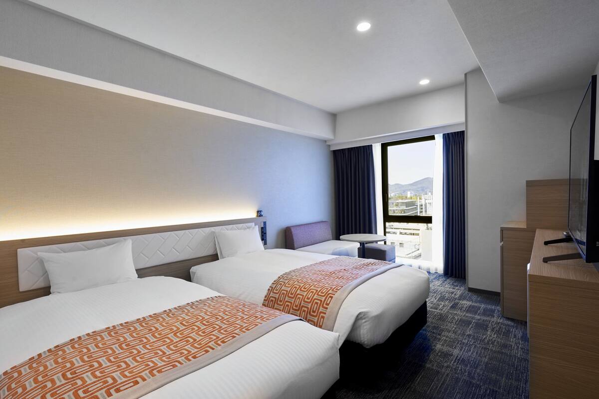 Habitación de hotel con dos camas, televisión, escritorio y balcón con vistas a la ciudad.
