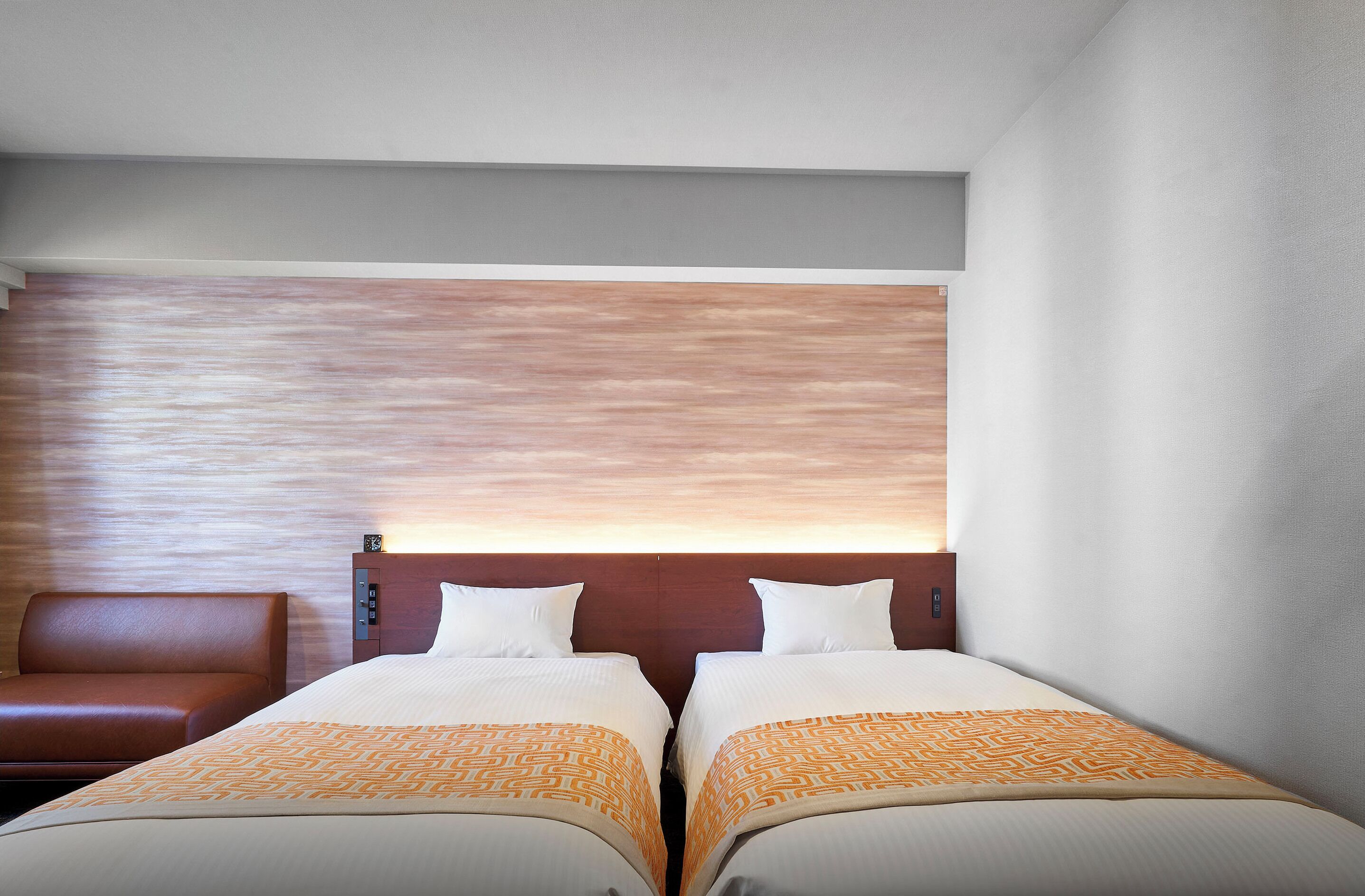 Une chambre d’hôtel avec deux lits, une tête de lit en bois, un fauteuil en cuir marron et une grande fenêtre donnant sur le ciel.