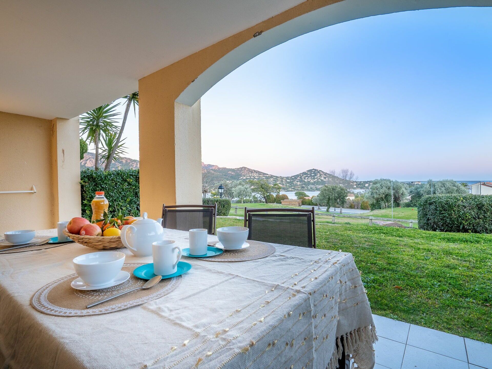 A dining table set with white tableware and a fruit basket, overlooking a lush garden and distant hills.