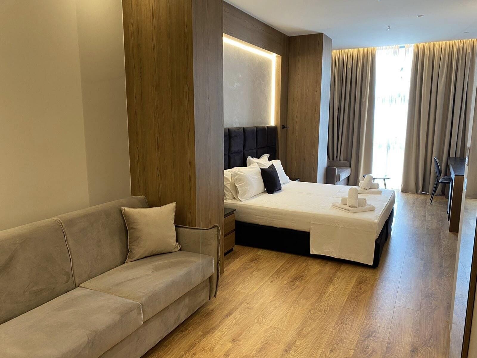 A modern hotel room with a large bed, a sofa, a desk, and a window with curtains.