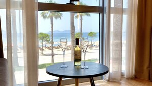 A round table with a bottle of wine and two glasses, positioned in front of a large window with sheer curtains, overlooking a beach with palm trees and a clear sky. - EL MAR HOTEL (Vlorë)