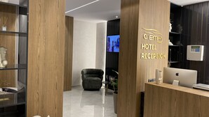 A hotel reception area with a wooden reception desk, a black armchair, and a television screen displaying a scene. - EL MAR HOTEL (Vlorë)