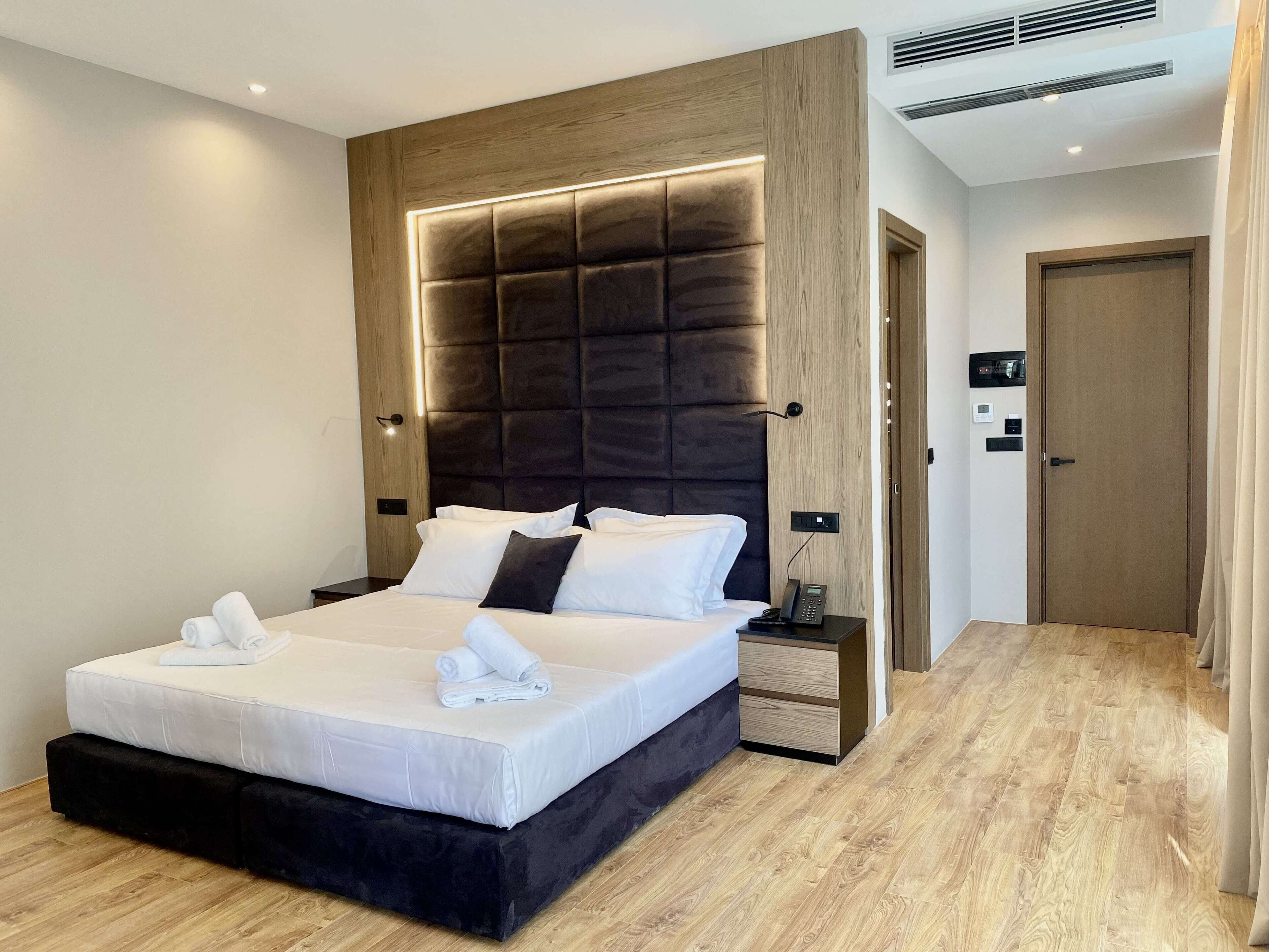 A modern hotel room with a large bed, a wooden headboard, a bedside table with a phone, and a door to the right.