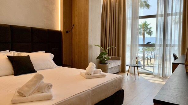 A hotel room with a large bed, a bedside table with a bottle and glass, a small table with a plant, and a view of the beach through large windows. - EL MAR HOTEL (Vlorë)
