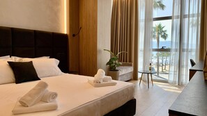 A hotel room with a large bed, a bedside table with a bottle and glass, a small table with a plant, and a view of the beach through large windows. - EL MAR HOTEL (Vlorë)
