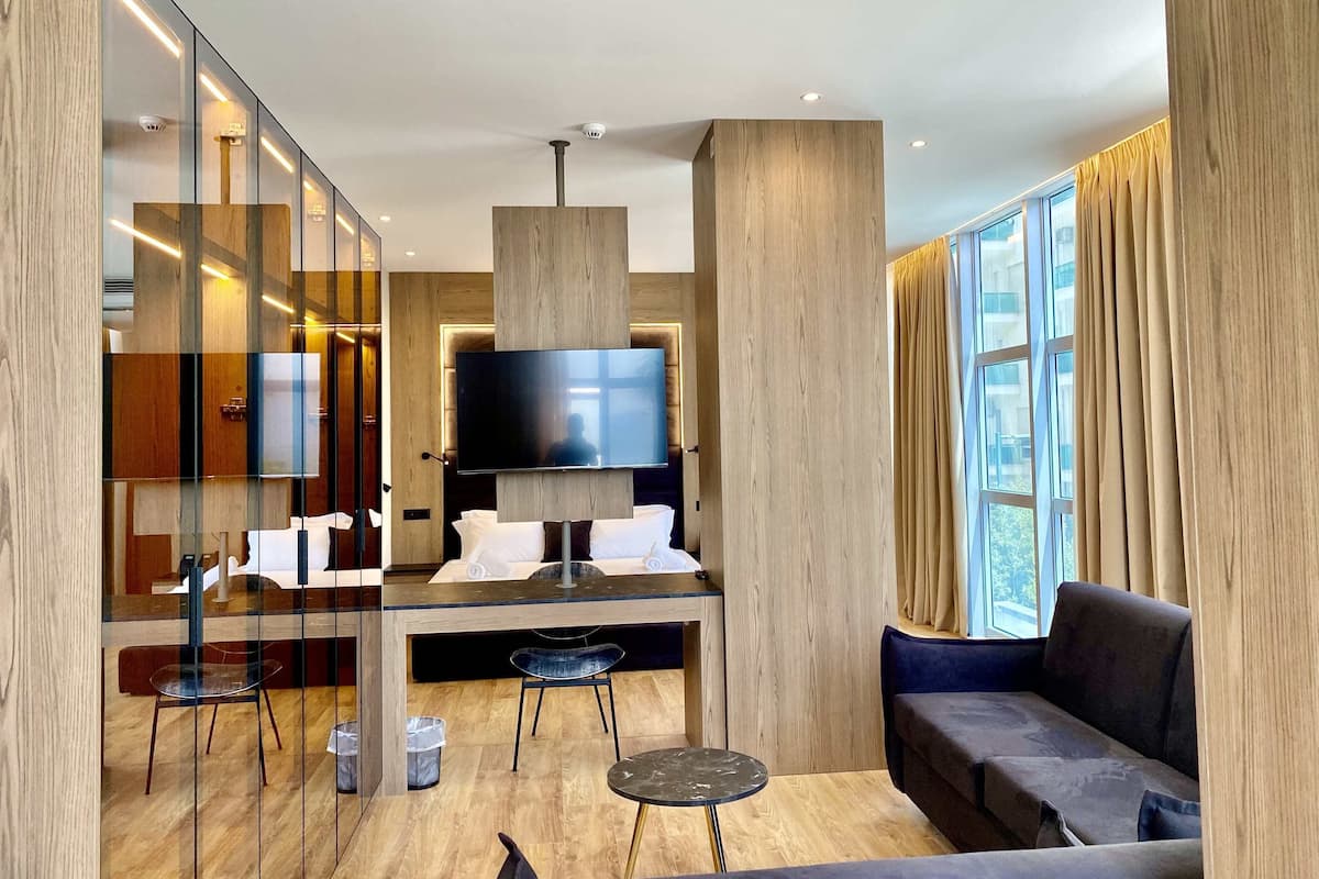 Une chambre d’hôtel moderne dotée d’une grande télévision à écran plat, d’un canapé, d’un coin repas avec une table et des chaises, et d’une grande fenêtre avec des rideaux.