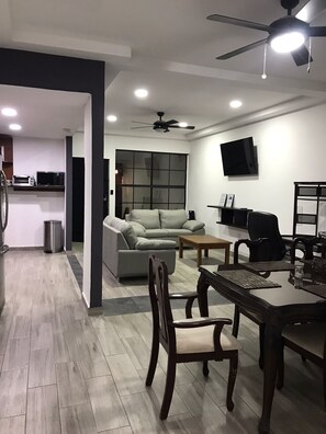 A modern living room with a sofa, dining table, and chairs, a kitchen area with appliances, and a large window.