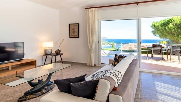 Una sala de estar moderna con televisor de pantalla plana, una mesa de centro de vidrio y un sofá con cojines estampados. Se ve una mesita auxiliar con una lámpara y objetos decorativos. La habitación tiene un amplio ventanal corredero que da a un espacio exterior con vistas al mar y un árbol.
