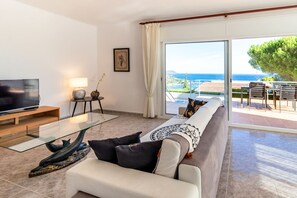 Uma sala de estar moderna com televisão de tela plana, mesa de centro de vidro e sofá com almofadas estampadas. Uma pequena mesa lateral com abajur e objetos decorativos é visível. O quarto possui uma grande porta de vidro deslizante que dá para uma área externa com vista para o mar e uma árvore.