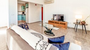 Una sala moderna con una mesa de centro de vidrio, un sofá con cojines estampados, un televisor sobre un mueble de madera, y una cocina visible a través de un vano abierto.