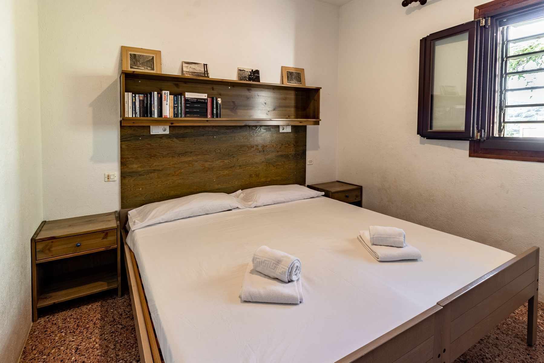 Un lit bien fait, avec deux serviettes, une tête de lit en bois, une table de chevet et une étagère avec des livres et des photos.