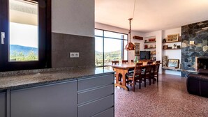 Een moderne keuken met granieten aanrechtbladen, een eethoek met een houten tafel en stoelen, een stenen openhaard en een groot raam met uitzicht op de omgeving.