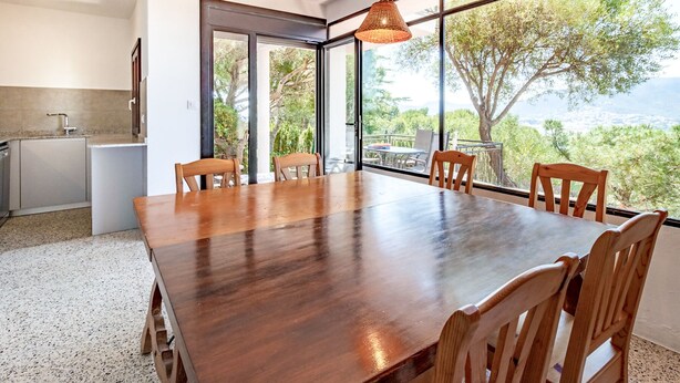 A dining area with a wooden table and chairs, large windows with a view of trees and hills, and a kitchen area with a sink and countertop. - Grifeu 08 - Apartment with outside terrace (Llança)