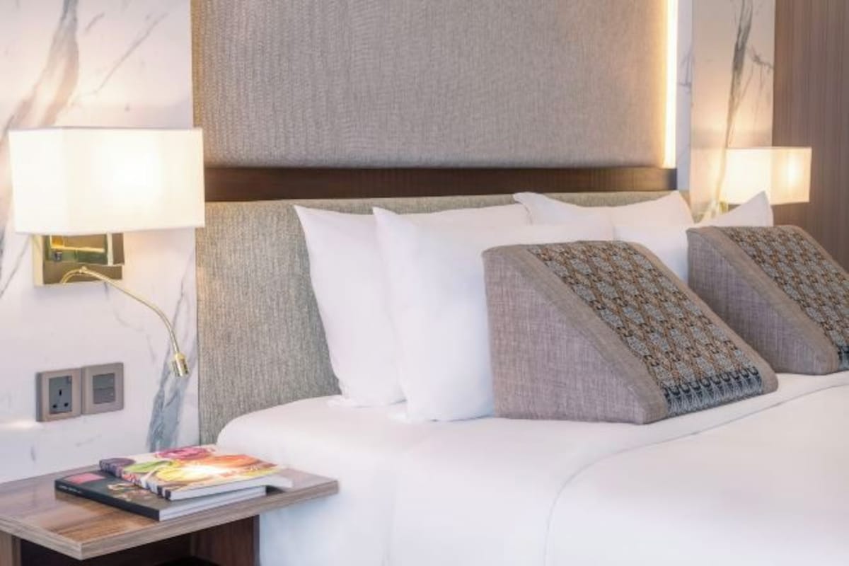 A hotel room with a large bed, two bedside lamps, a nightstand with a book, and a wall-mounted light fixture.