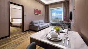 A hotel room with a bed, a sofa, a dining area with a table set for two, and a large window offering a city view.