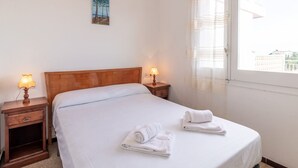 Una cama bien hecha con sábanas blancas y dos toallas enrolladas. Hay dos mesitas de noche con lámparas, un cabecero de madera y una ventana con cortinas translúcidas.