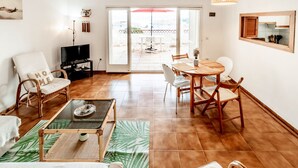 Amplia sala de estar con televisor, una mesa de comedor con sillas, una silla de mimbre con cojín y un gran ventanal corredero que da a un patio exterior.