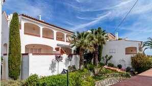 Un edificio blanco con varios balcones, un sendero de piedra y abundante vegetación, incluyendo palmeras.