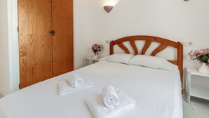 A neatly made bed with white linens and two rolled towels, a wooden headboard, a nightstand with a vase of flowers, and a wooden door. - Sant Carles 25 - apartment with terrace (Llança)