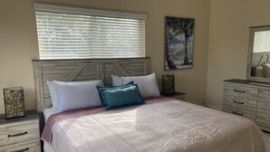 A bedroom with a wooden headboard, a large bed with patterned bedding, a window with blinds, a painting on the wall, and a dresser with drawers. - Key Largo New Construction 2-story! Exclusive Rights for 28 Days! (Key Largo)