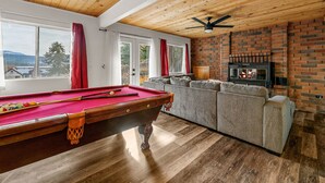 Ein Billardtisch mit Kugeln, eine Couch und ein Kamin in einem Raum mit Holzbalkendecken und großen Fenstern.