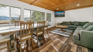 Ein geräumiges Wohnzimmer mit Holzboden, großen Fenstern, einem grünen Sofa und einem Essbereich mit Holzmöbeln.