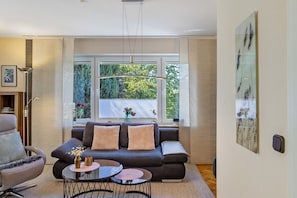 A modern living room with a sofa, two side tables, a chair, a large window with blinds, and a hanging light fixture.