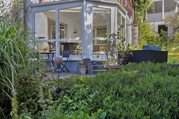 A glass-enclosed patio with outdoor furniture, surrounded by lush greenery.
