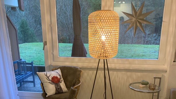 Ett rum med ett stort fönster, en golvlampa, en stol med en kudde och ett litet bord med ett glas på.