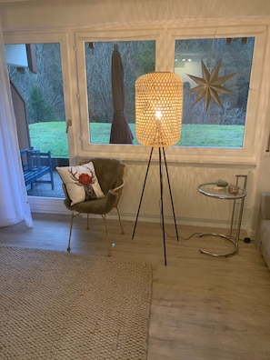 Una habitación con un ventanal grande, una lámpara de pie, una silla con cojín y una mesita con un vaso.