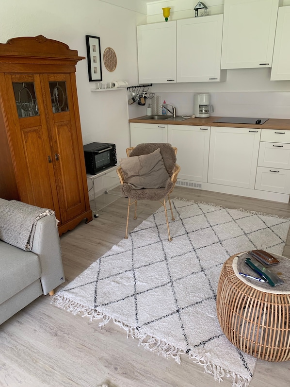 Una sala de estar con un sofá, un aparador de madera, una silla de mimbre y una zona de cocina con armarios blancos y un microondas.