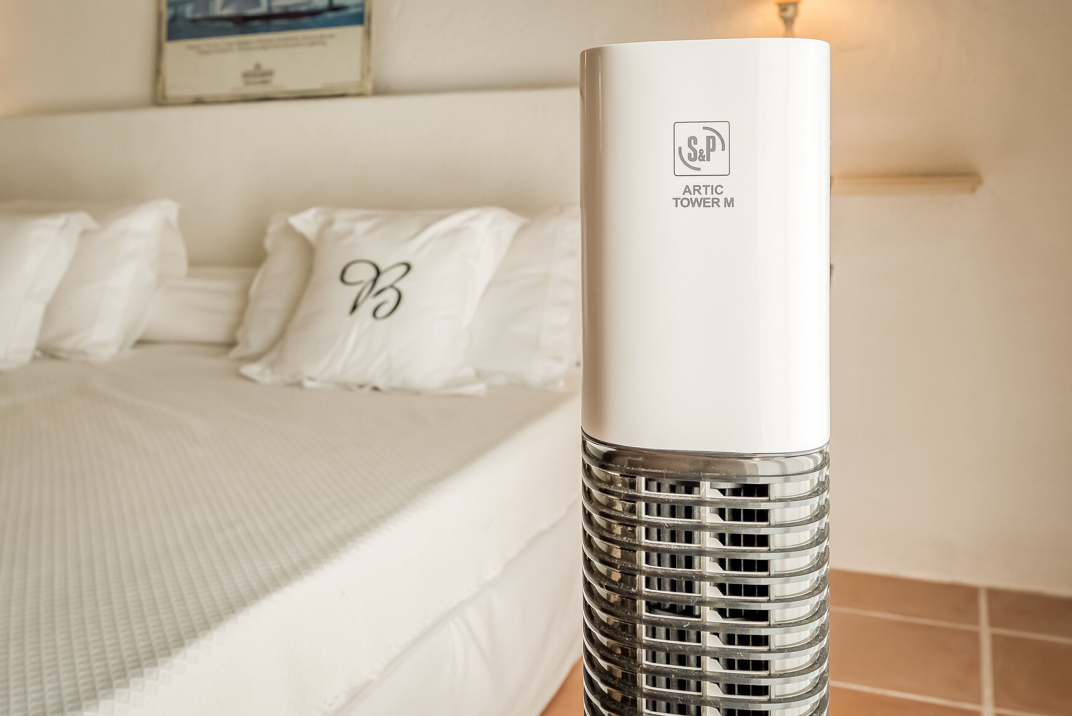 Ein hoher, zylindrischer Luftreiniger mit Metallgitter und einem Logo an der Seite, platziert in einem Hotelzimmer mit einem ordentlich bezogenen Bett und weißen Kissen.