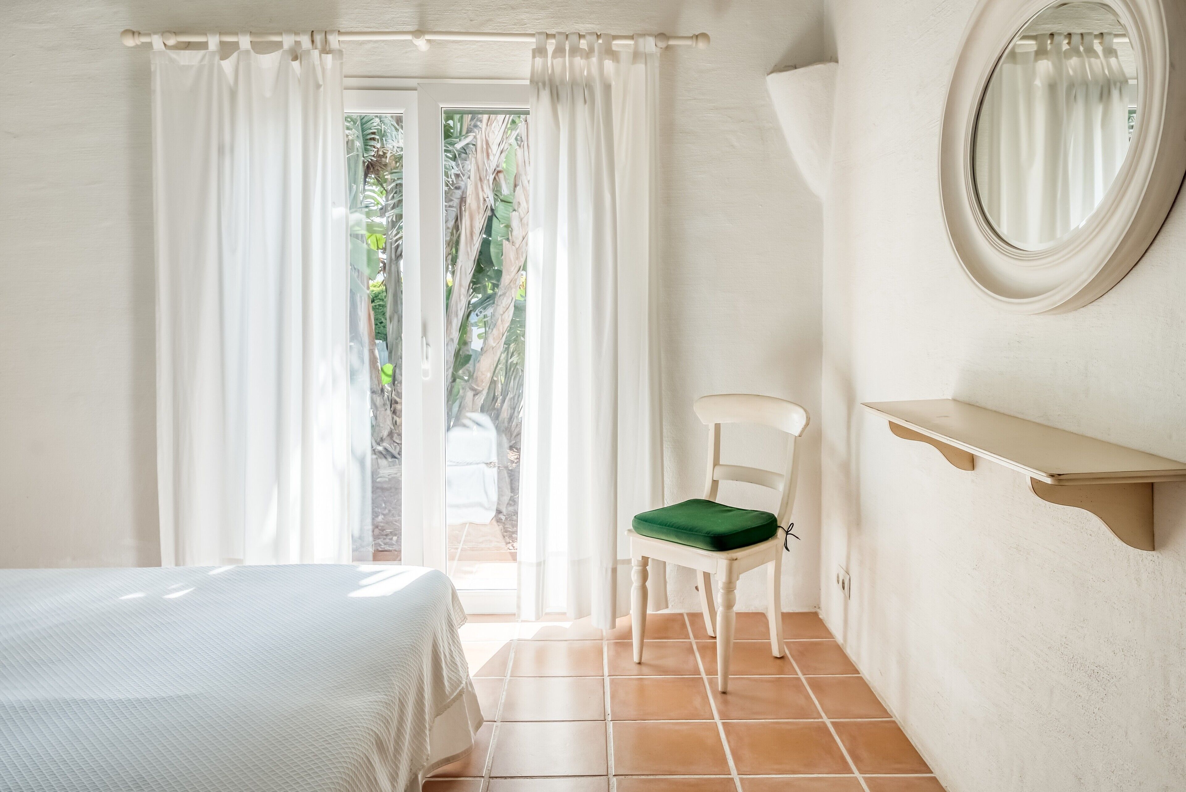 A bedroom with a bed, a chair with a green cushion, a small table, a mirror, and a view through a window with curtains.