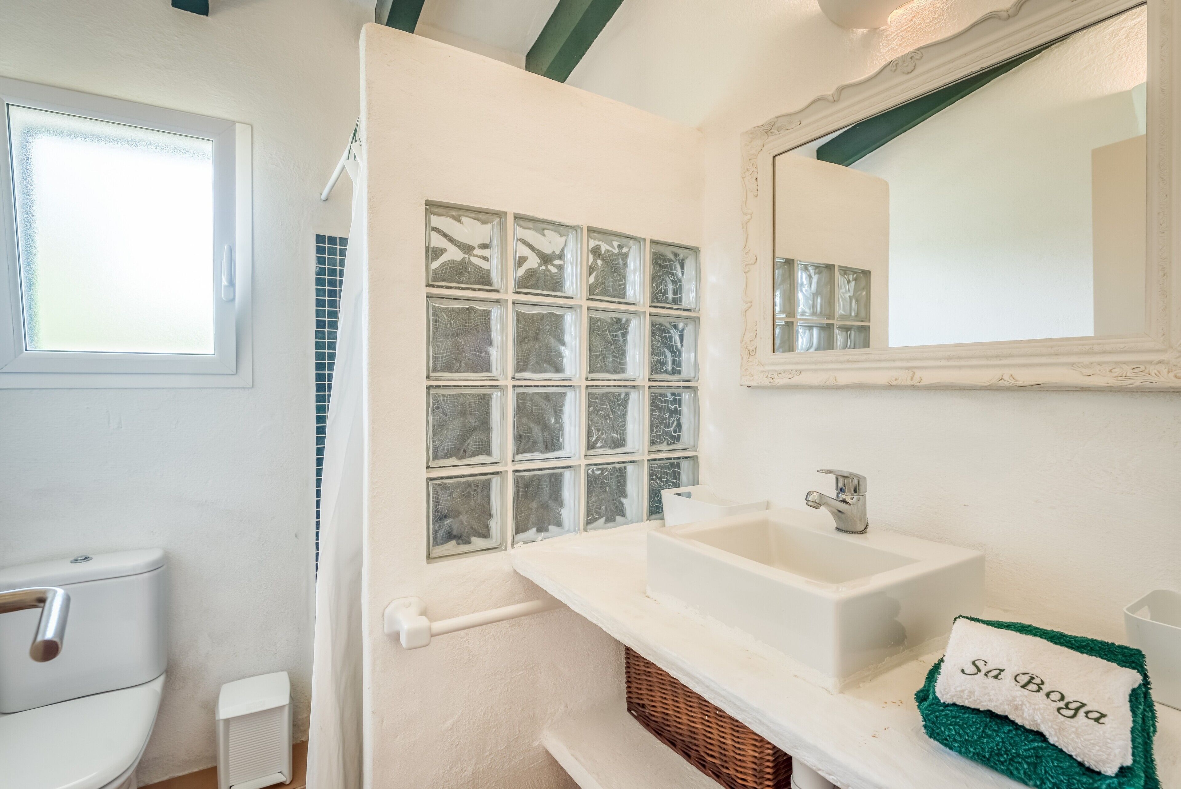 Un baño con lavamanos blanco, un espejo, una ventana con vidrio esmerilado y una toalla con la inscripción 'Sa Boja'.