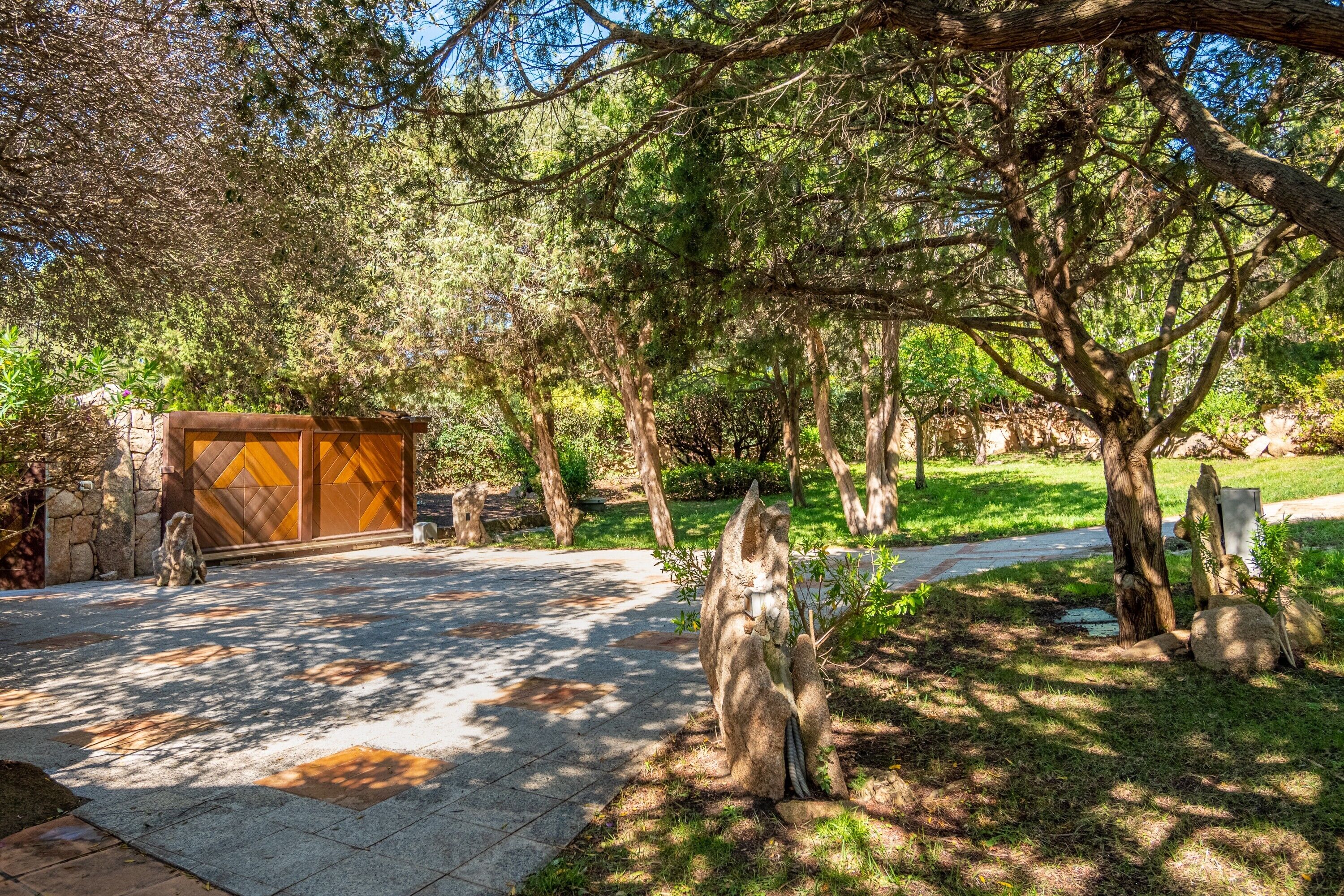 A paved pathway leading to a wooden gate, surrounded by trees and a stone wall.