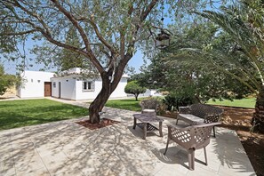 Un patio con mobiliario exterior, un árbol y un edificio blanco al fondo.