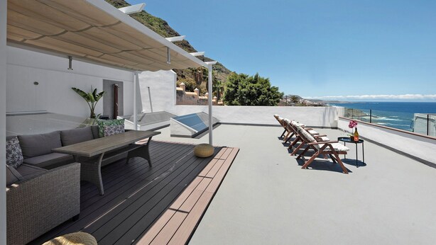 Une terrasse sur le toit, aménagée avec un ensemble de canapés, une table et des chaises longues, offrant une vue sur un paysage côtier.